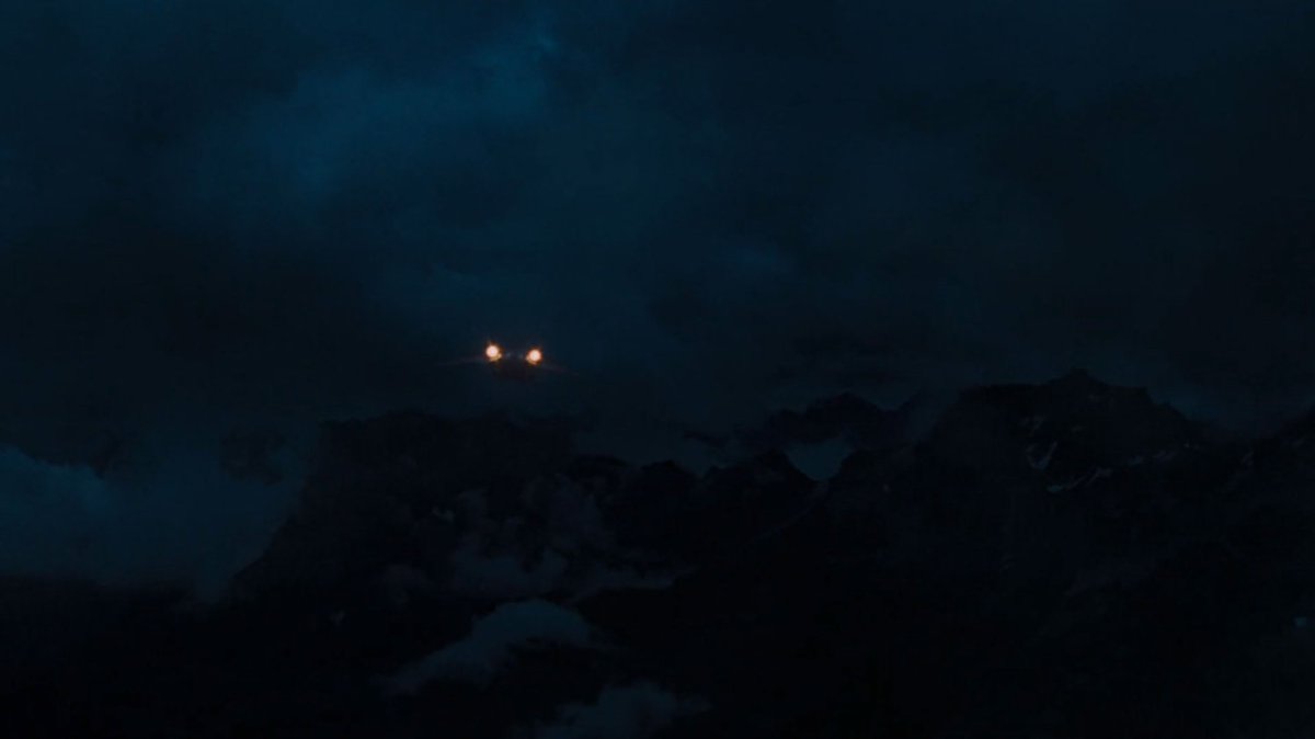 A quinjet approaches a dark mountain range.